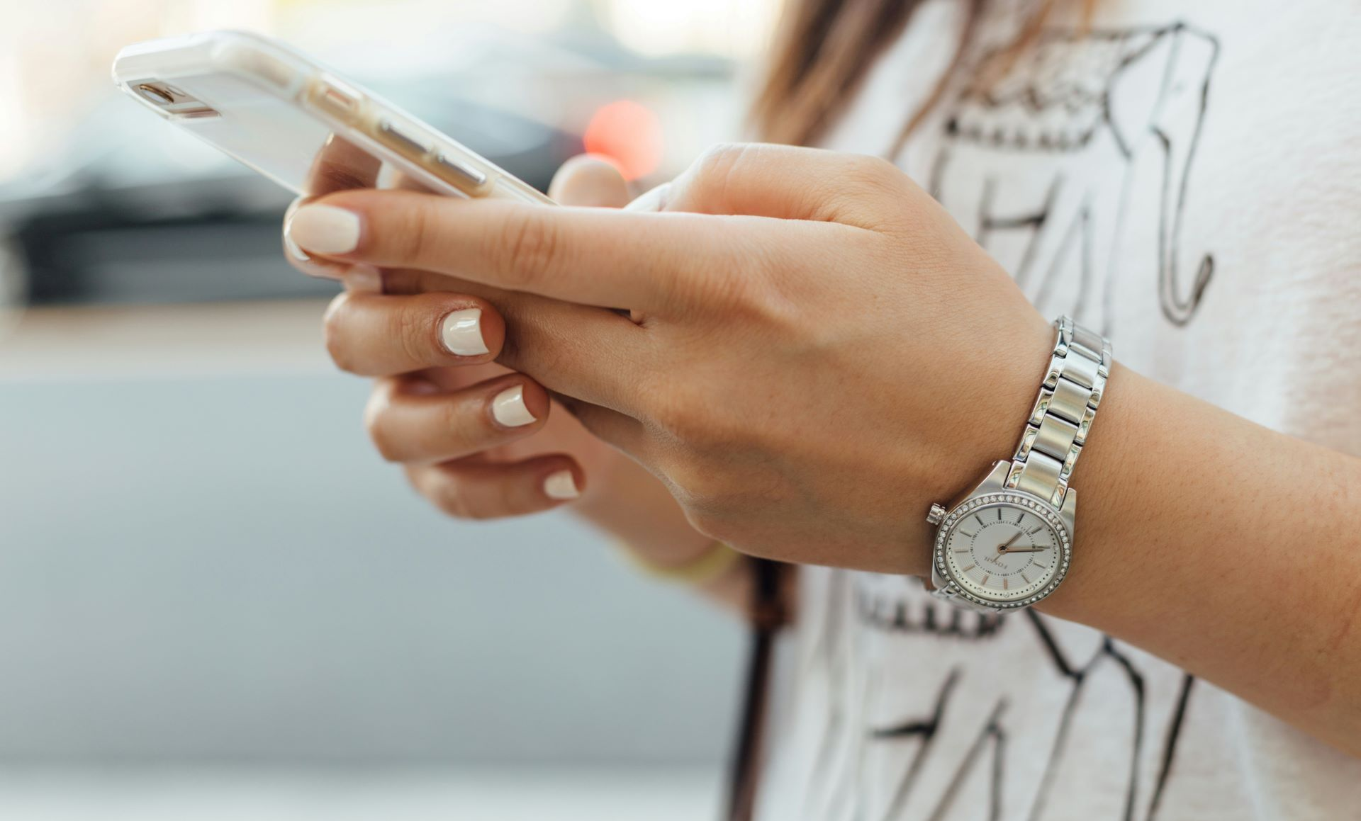 Hands holding a smartphone with a silver watch and white manicure in a bright indoor setting
