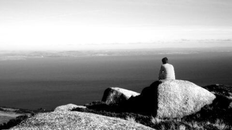 Person sits on a large rock overlooking a calm ocean under a bright sky.