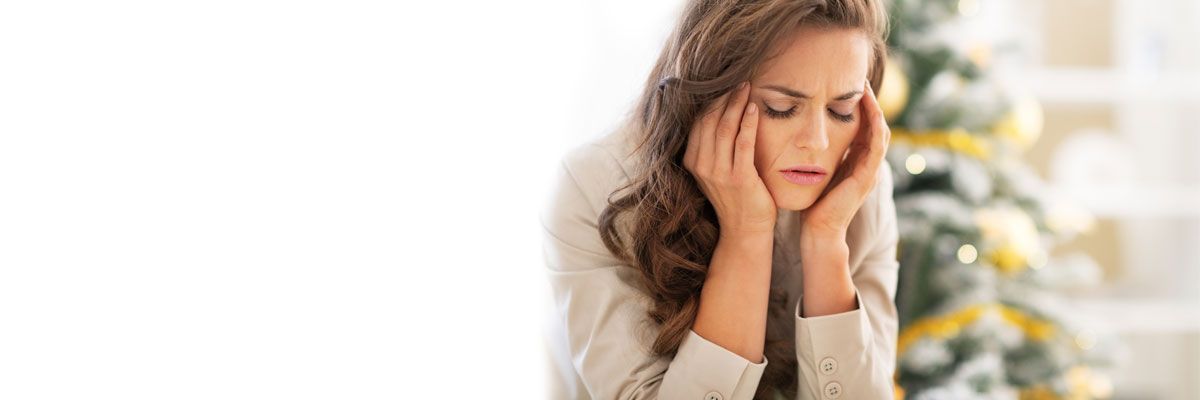 Woman holding her head in pain beside a softly lit Christmas tree