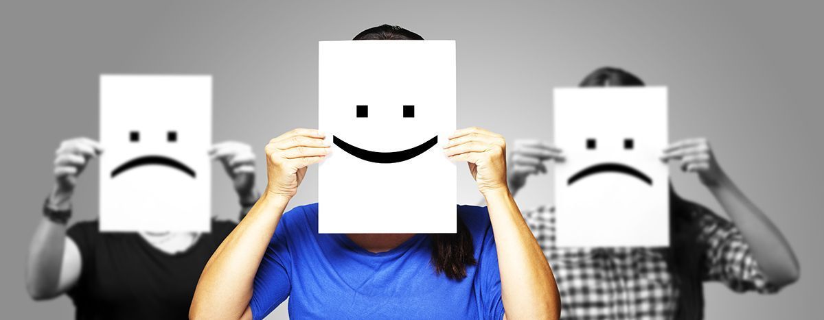 Three people holding sad-face paper masks, with one smiling person in a blue shirt in the center