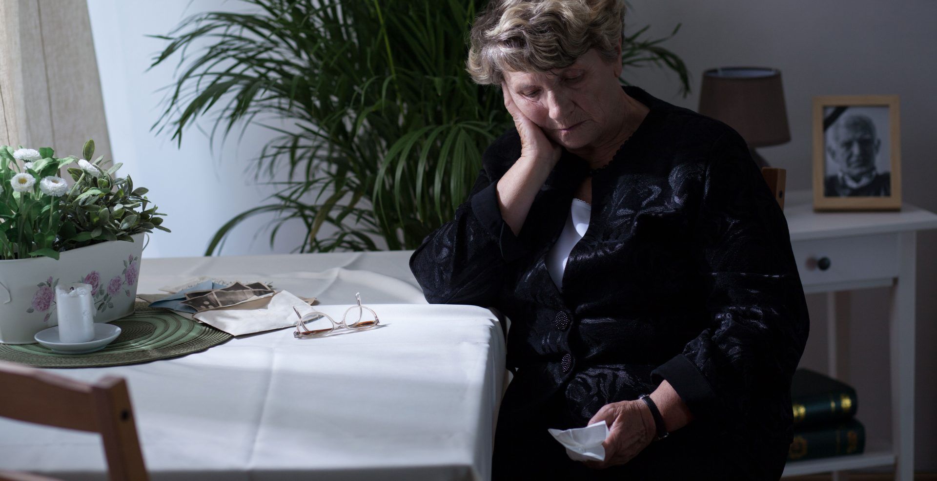 Person in black clothes sits at a white table, resting their head on one hand in a bright room.
