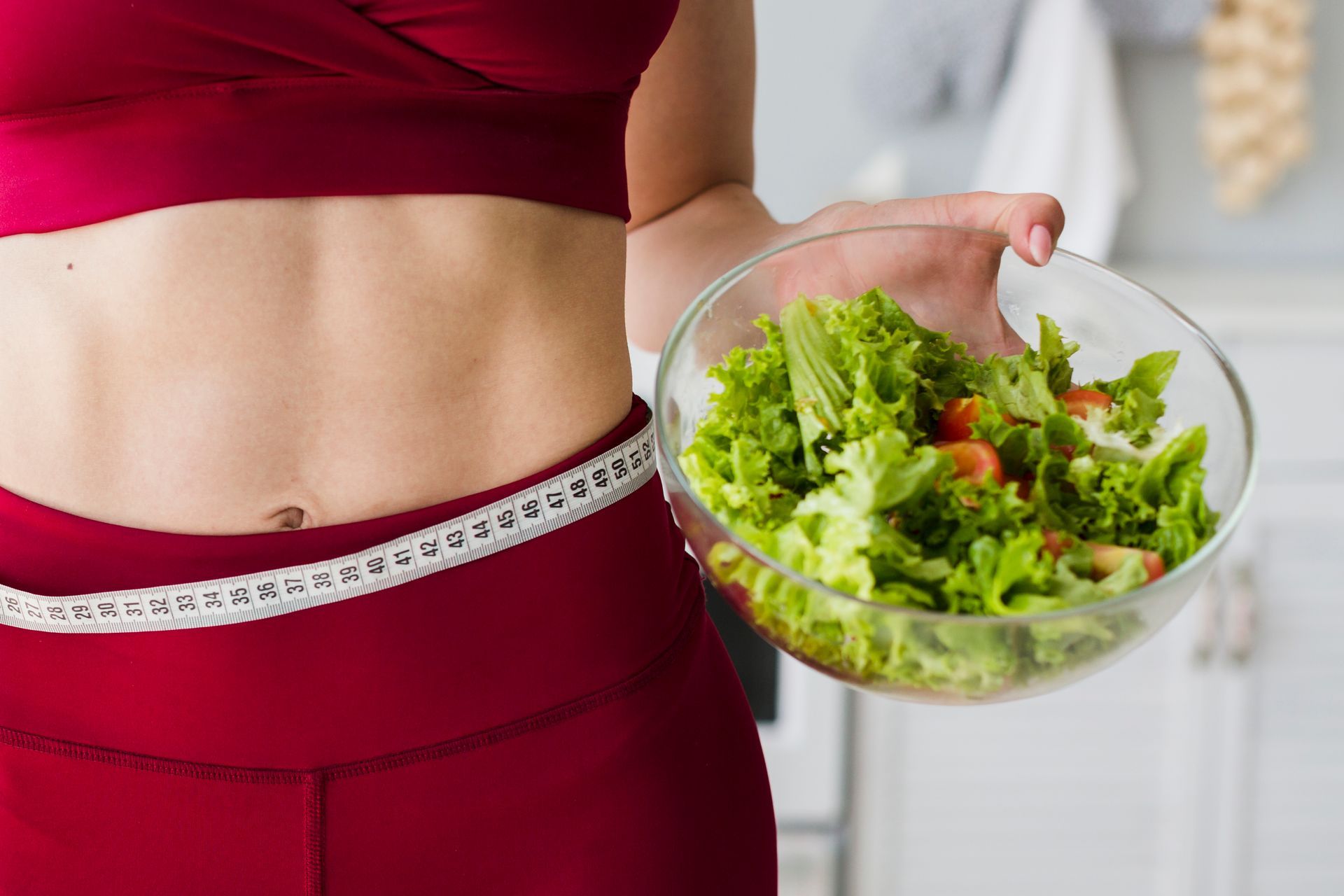 A woman is measuring her waist while holding a bowl of salad.
