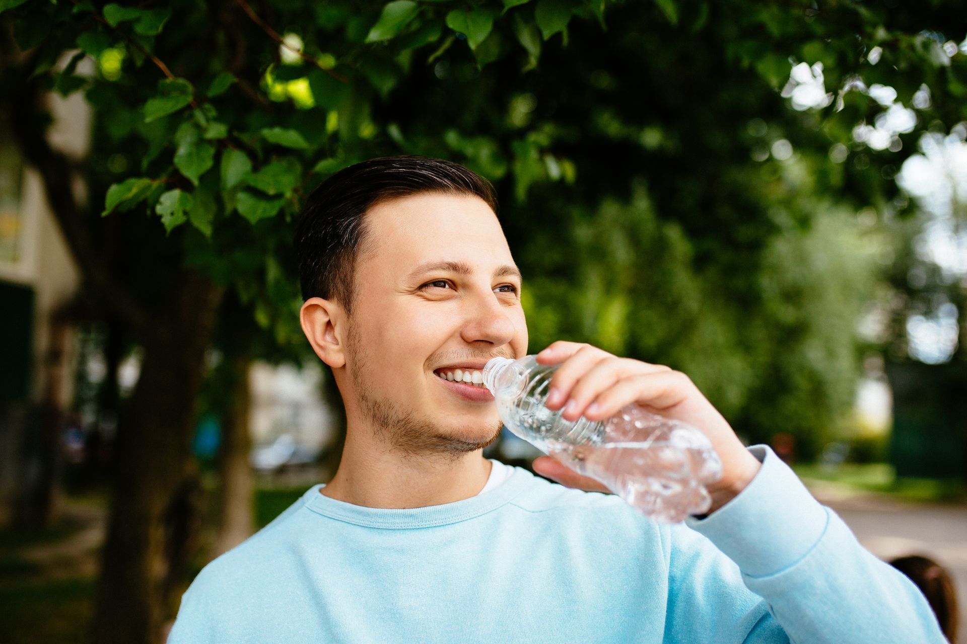 A young man is drinking water from a plastic bottle in a park.