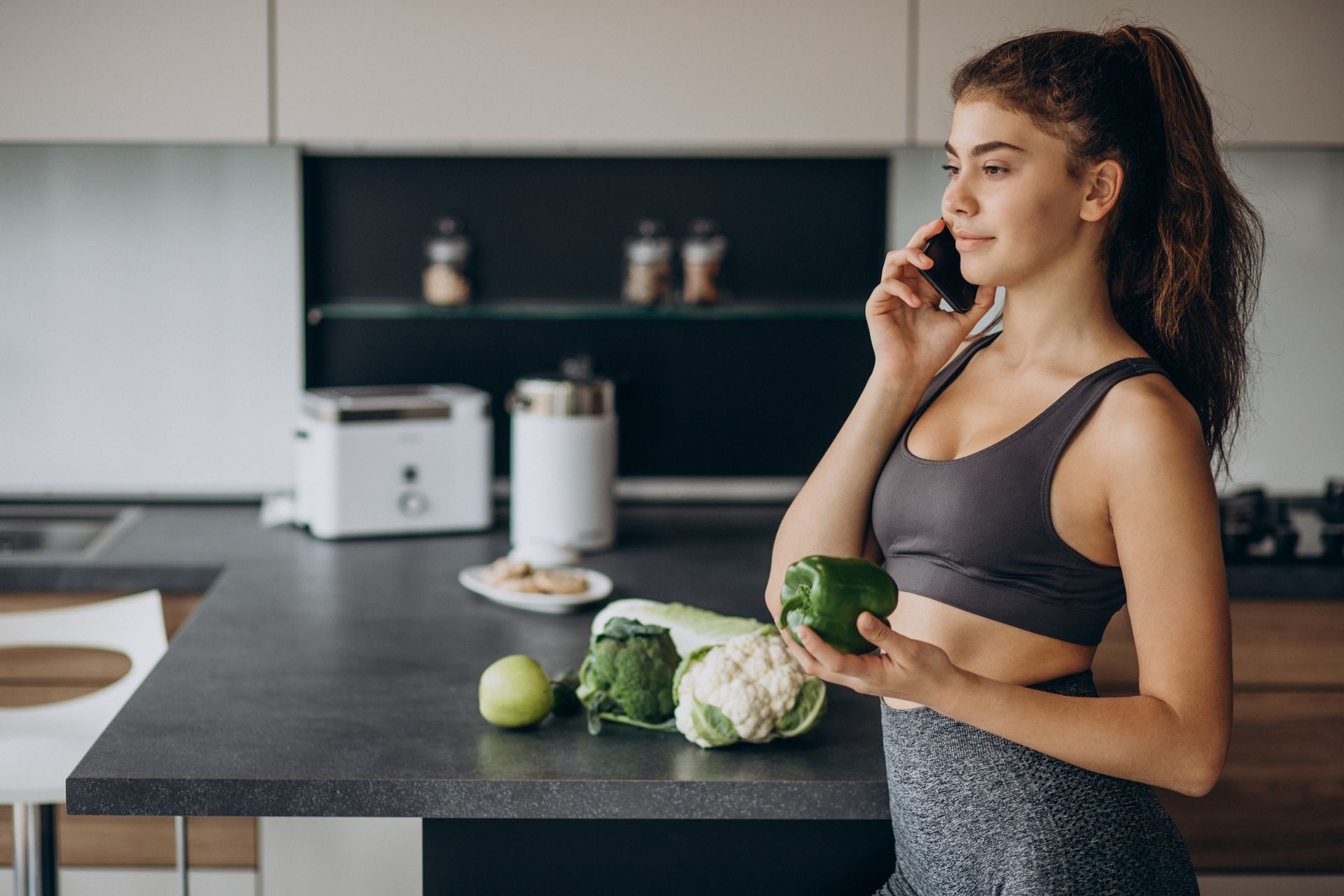 A woman is talking on a cell phone while holding a pepper in a kitchen.