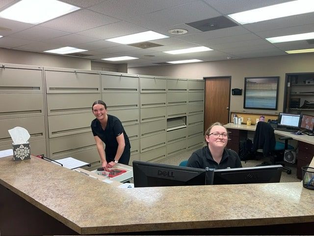 Two smiling office workers at a reception desk with computer monitors, file cabinets, and artwork.