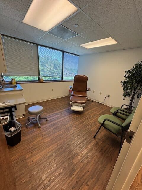 Medical exam room with examination chair, stool, chairs, and a small tree. Windows with shades, wood floor.
