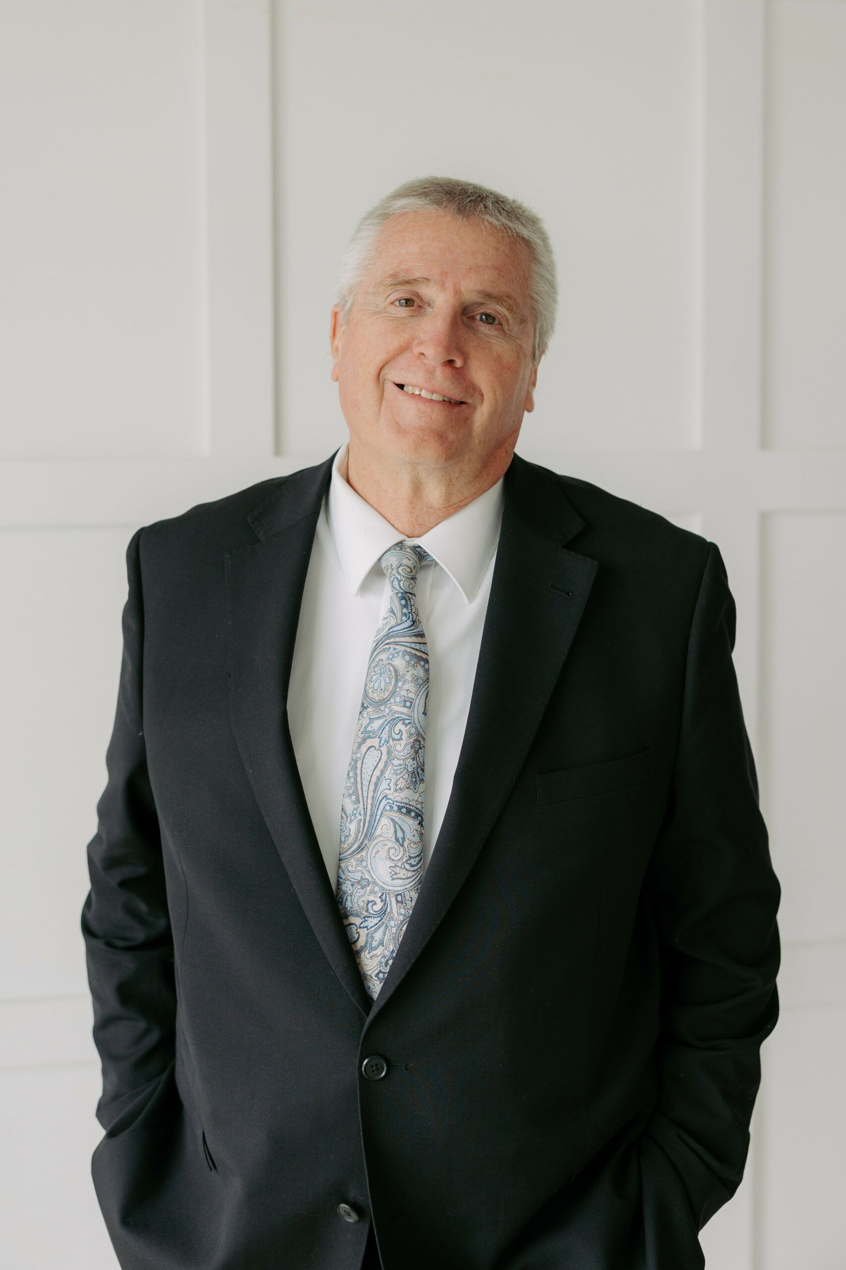 Smiling man in a dark suit and patterned tie standing against a light wall