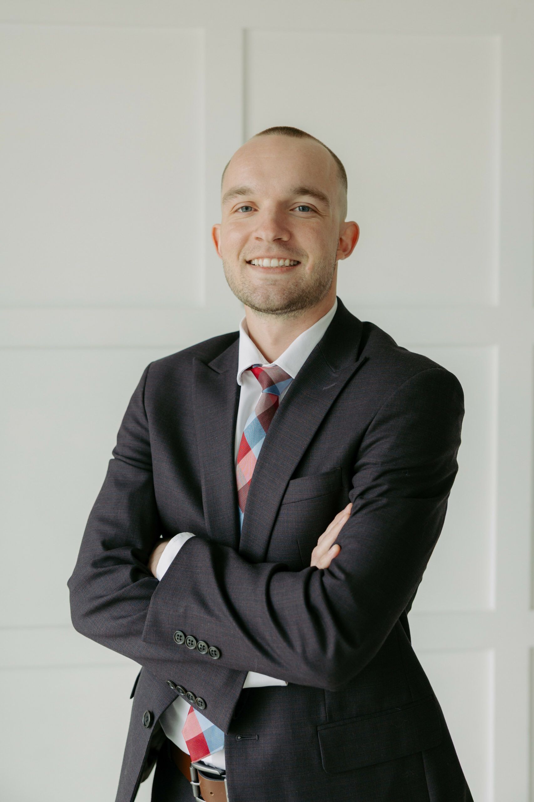 Smiling man in a dark suit with a striped tie, standing with arms crossed against a white wall