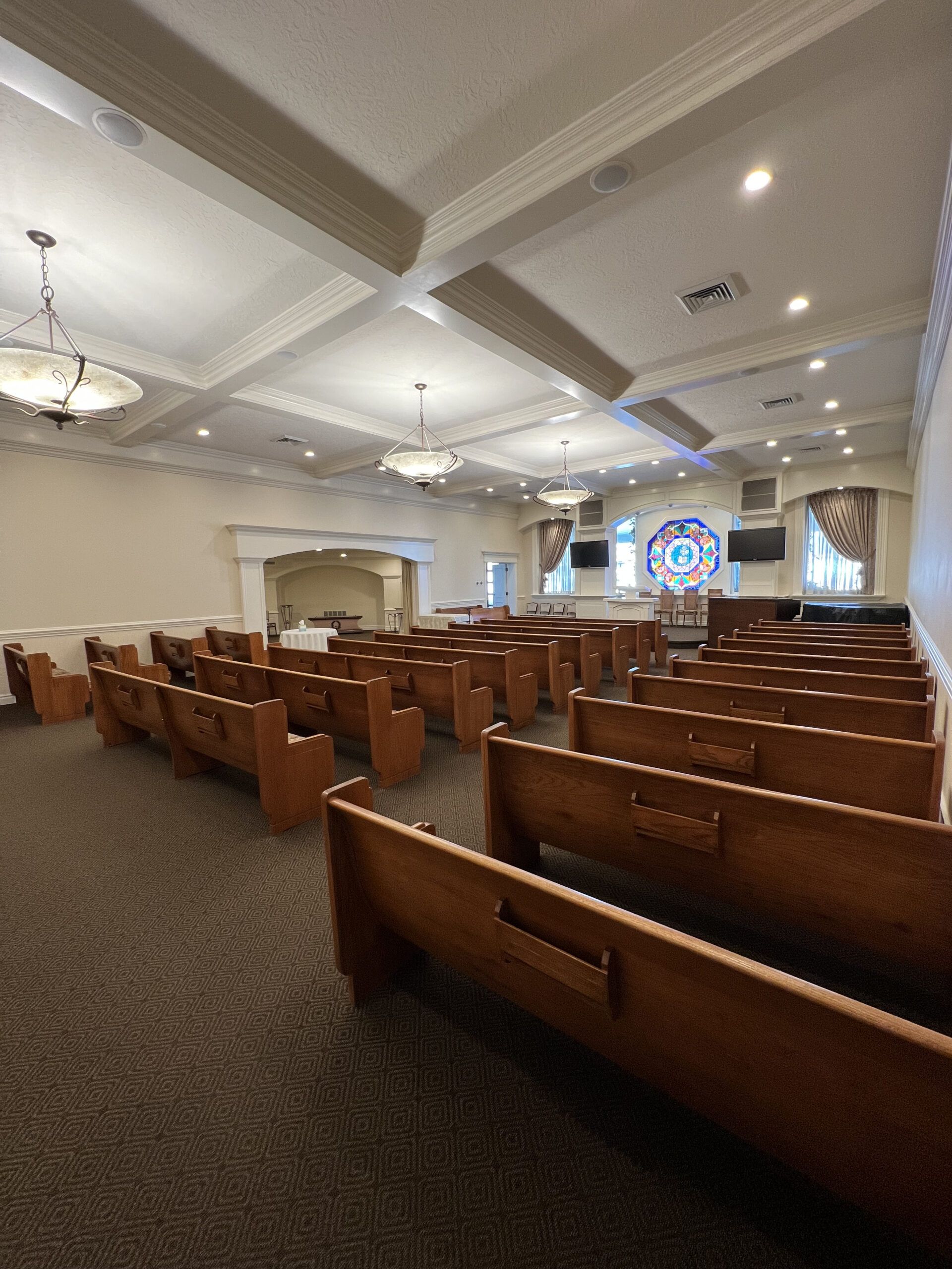 Empty church sanctuary with wooden pews, white coffered ceiling, and stained-glass window at the front.