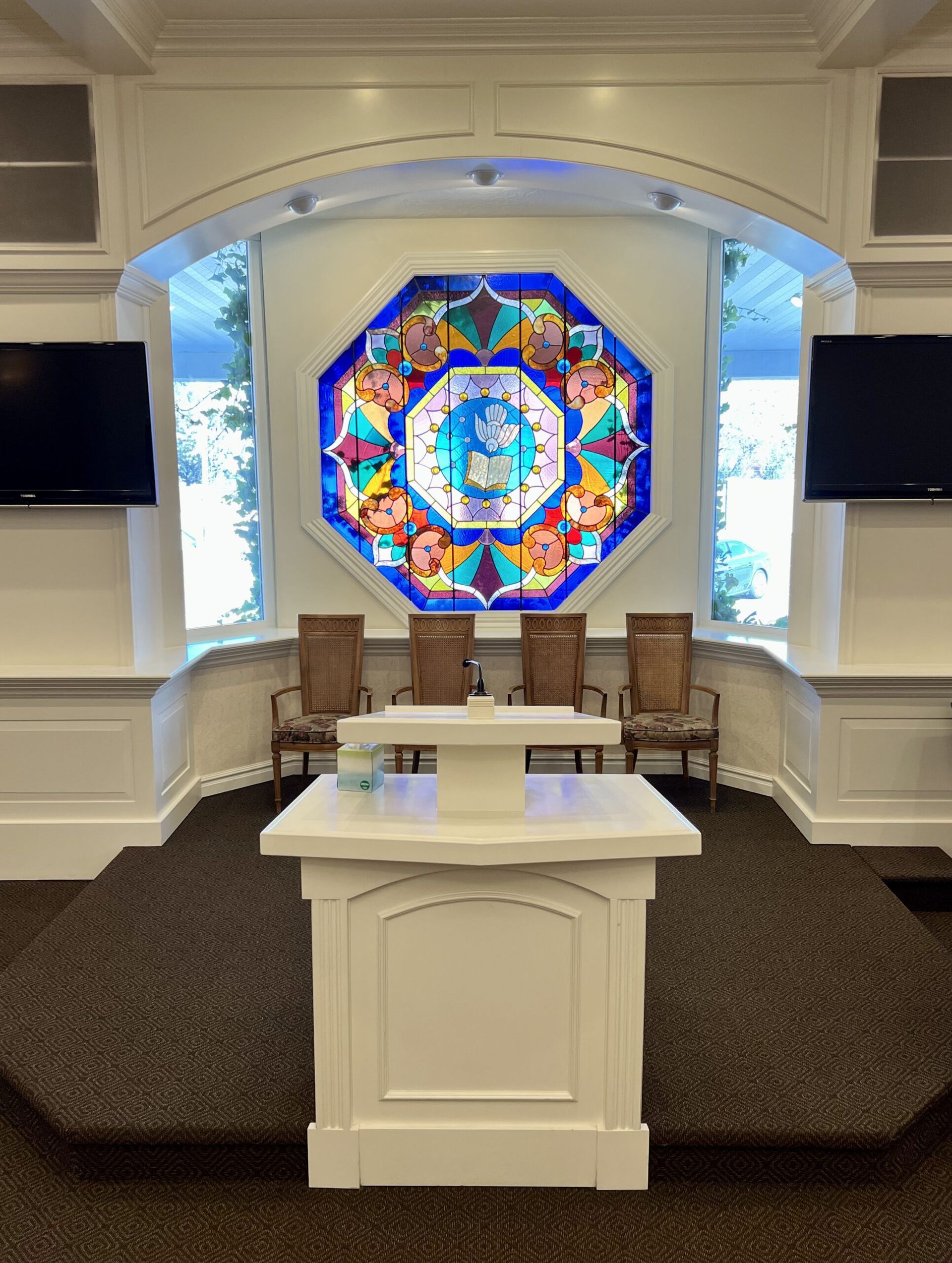 White chapel interior with stained-glass window, altar, and chairs on a raised platform