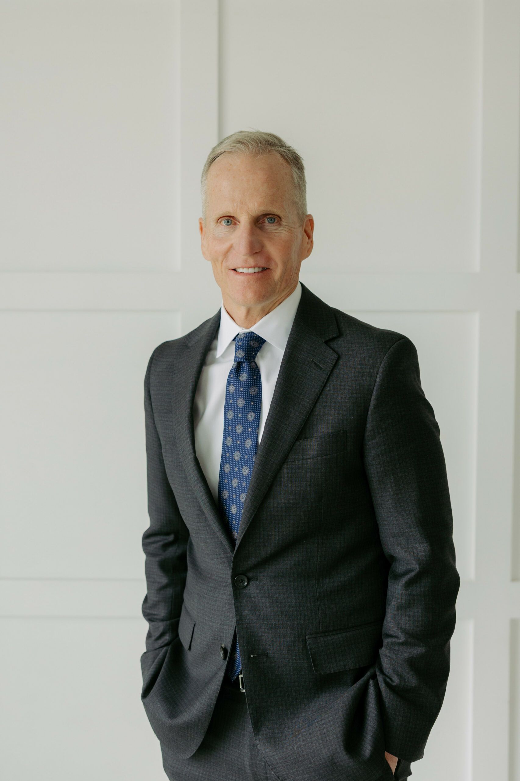Man in a dark suit and blue tie standing indoors with hands in pockets, smiling at the camera