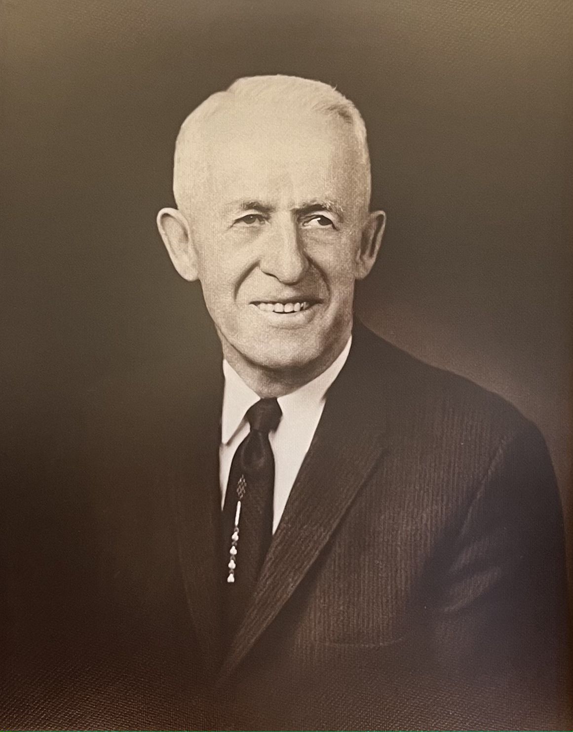 Studio portrait of a smiling man in a dark suit and tie against a plain brown background