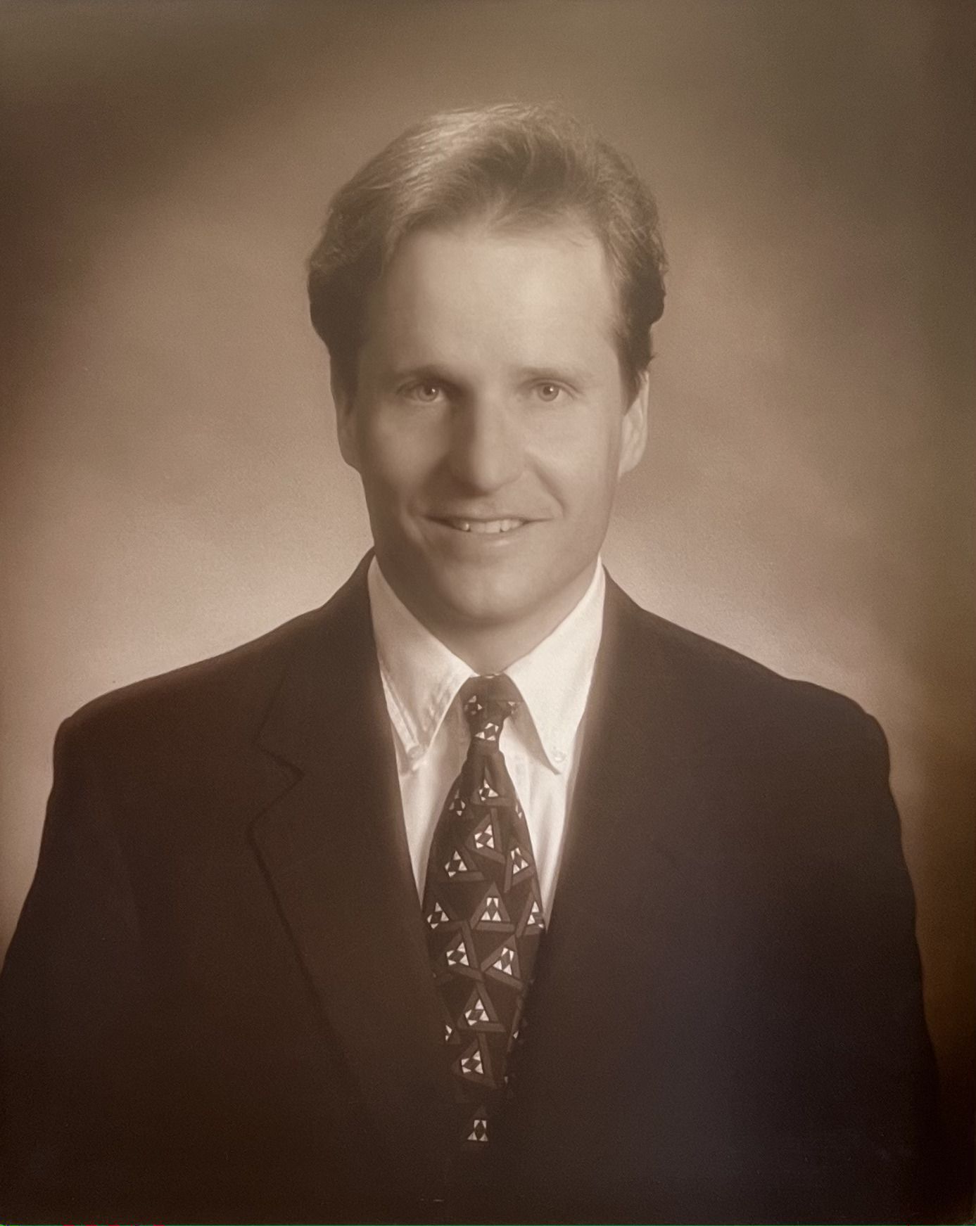 Studio portrait of a smiling man in a dark suit and patterned tie against a soft sepia background
