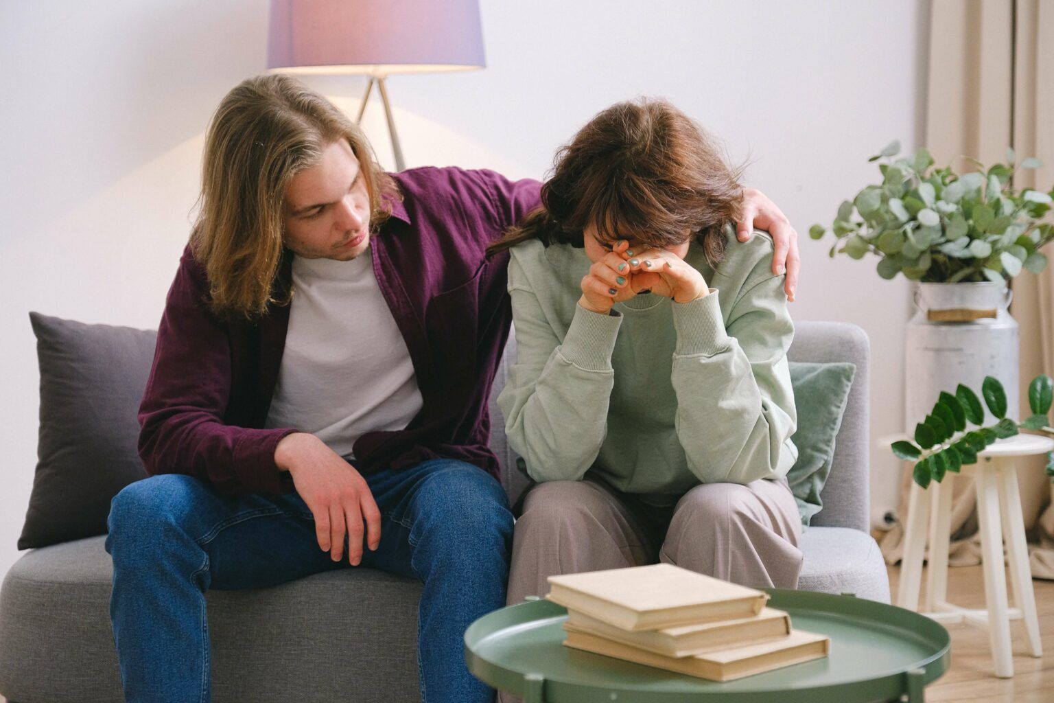 Two people sit on a couch, one consoling the other who is crying, with a coffee table and books nearby.