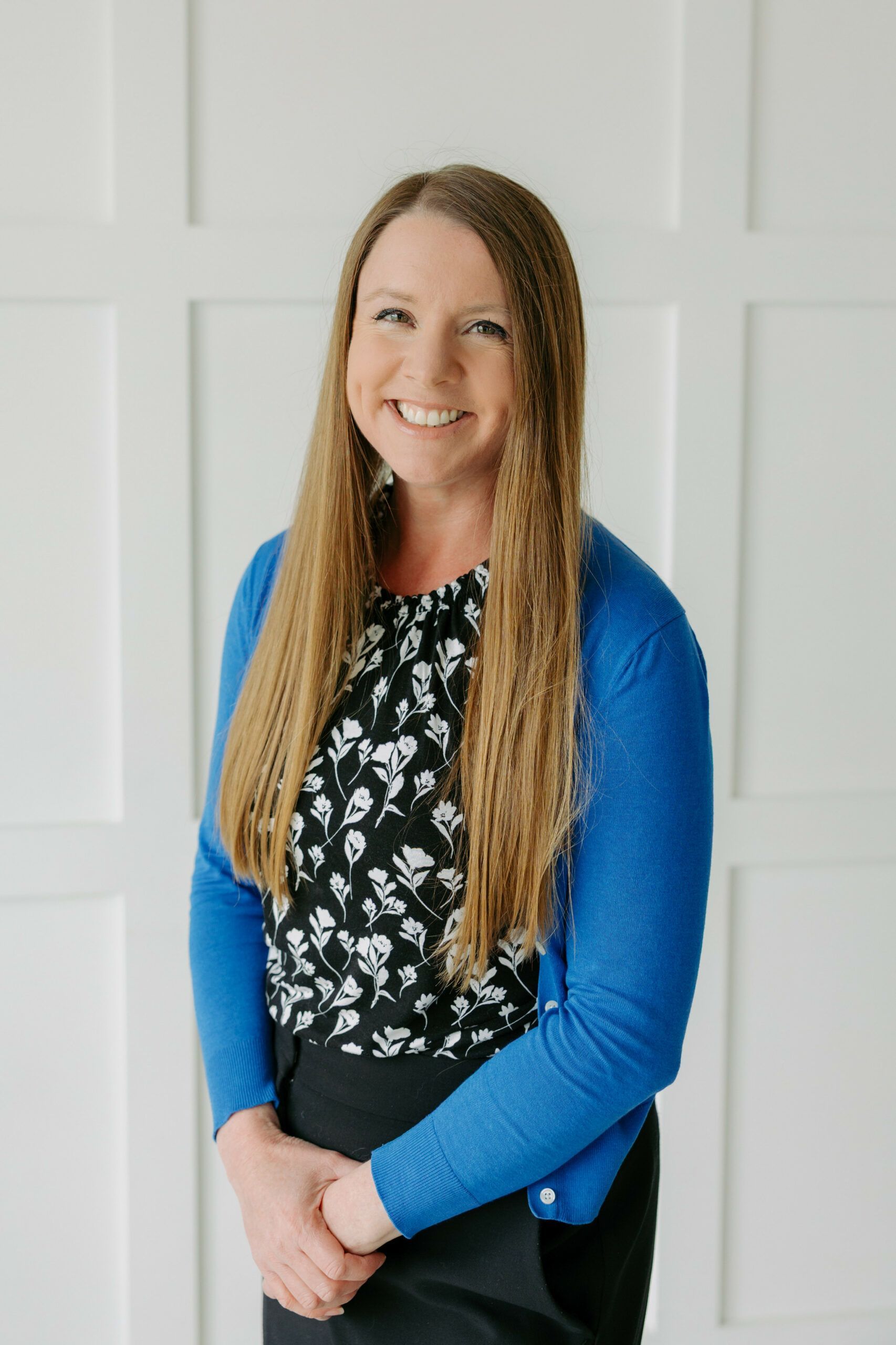 Smiling person in a blue cardigan over a black floral top, standing against a white paneled wall.