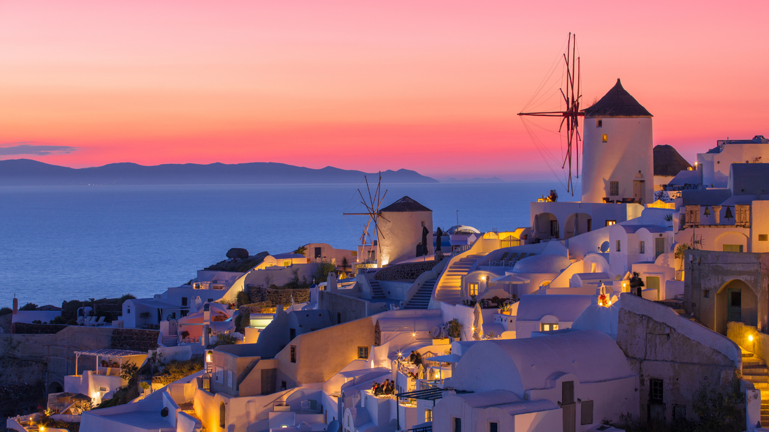 A sunset over the white buildings and iconic windmills of Oia, Santorini, with a pink sky reflecting on the sea.