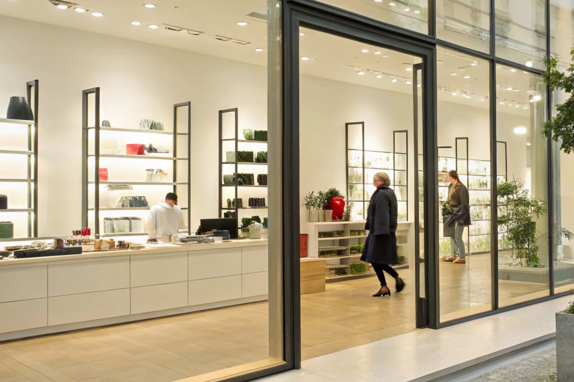 Shop interior with glass windows and a person entering. Shelves display products; a person works at a counter.
