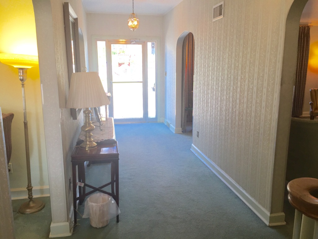 Hallway with blue carpet, beige walls, a side table lamp, and a bright doorway at the end