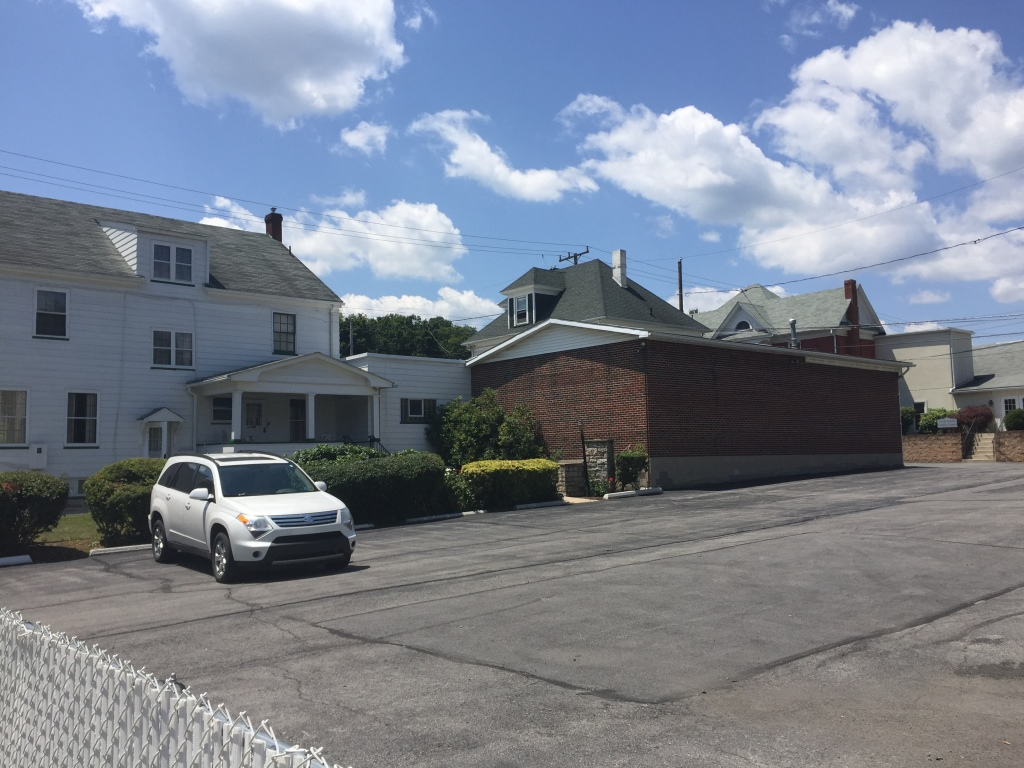 Parking lot with a white SUV beside a white building and brick structure under a partly cloudy sky