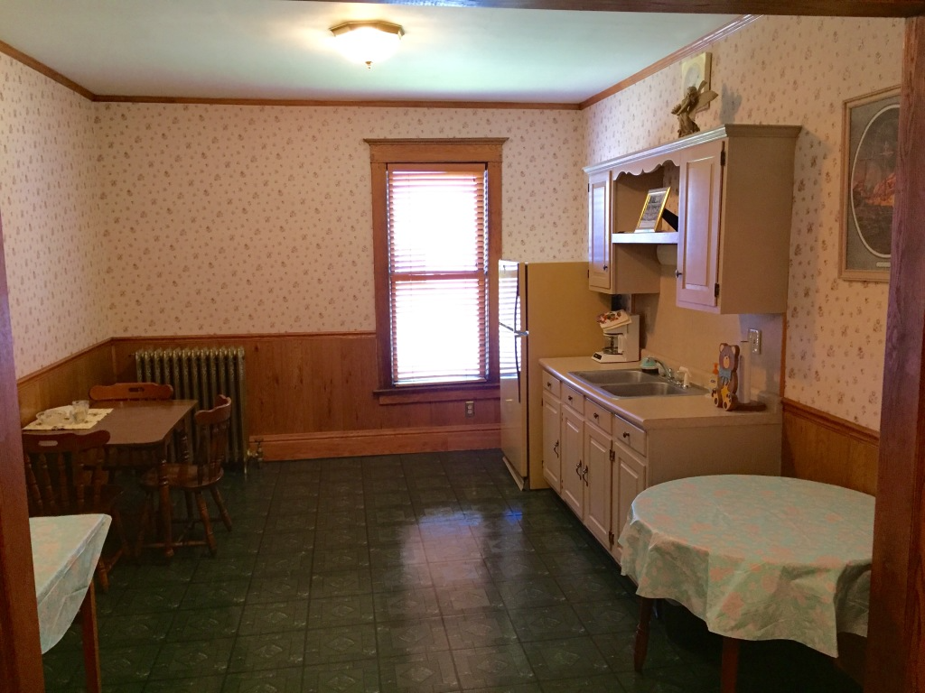 Small kitchen with wood cabinets, green floor, table, sink, window, and wallpapered walls.