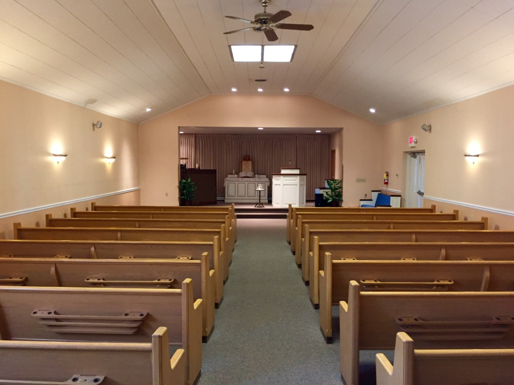 Empty church sanctuary with wooden pews, central aisle, stage, and ceiling fan under bright lights
