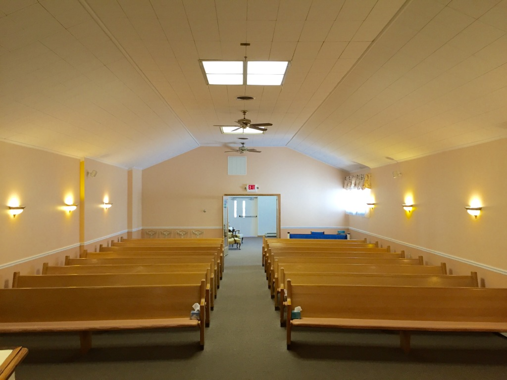 Empty church sanctuary with wooden pews, beige walls, wall sconces, and a ceiling fan near the front door