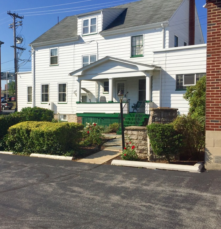 White two-story house with green porch and shrubs beside a paved street under a clear sky