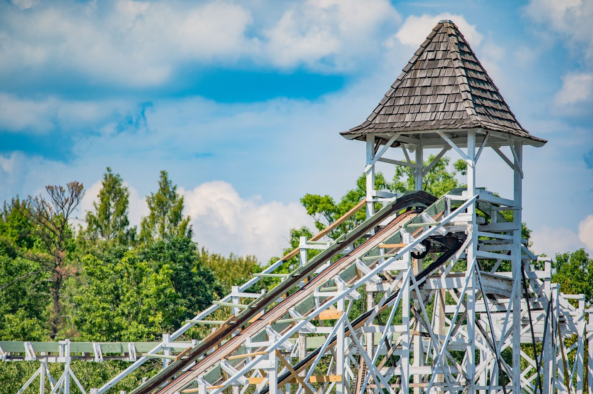 Wooden roller coaster with a shingled lift hill tower under a blue sky and trees