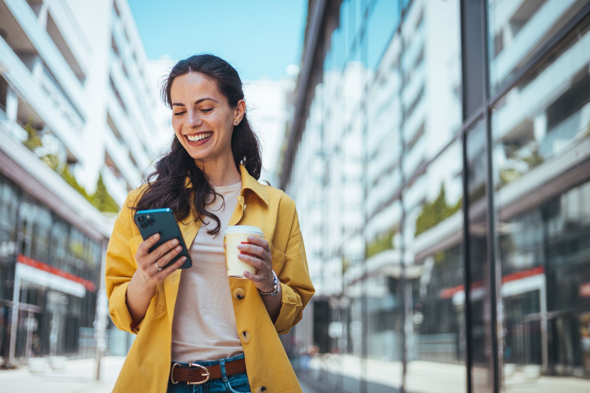 A woman in a yellow jacket is holding a cup of coffee and looking at her phone.