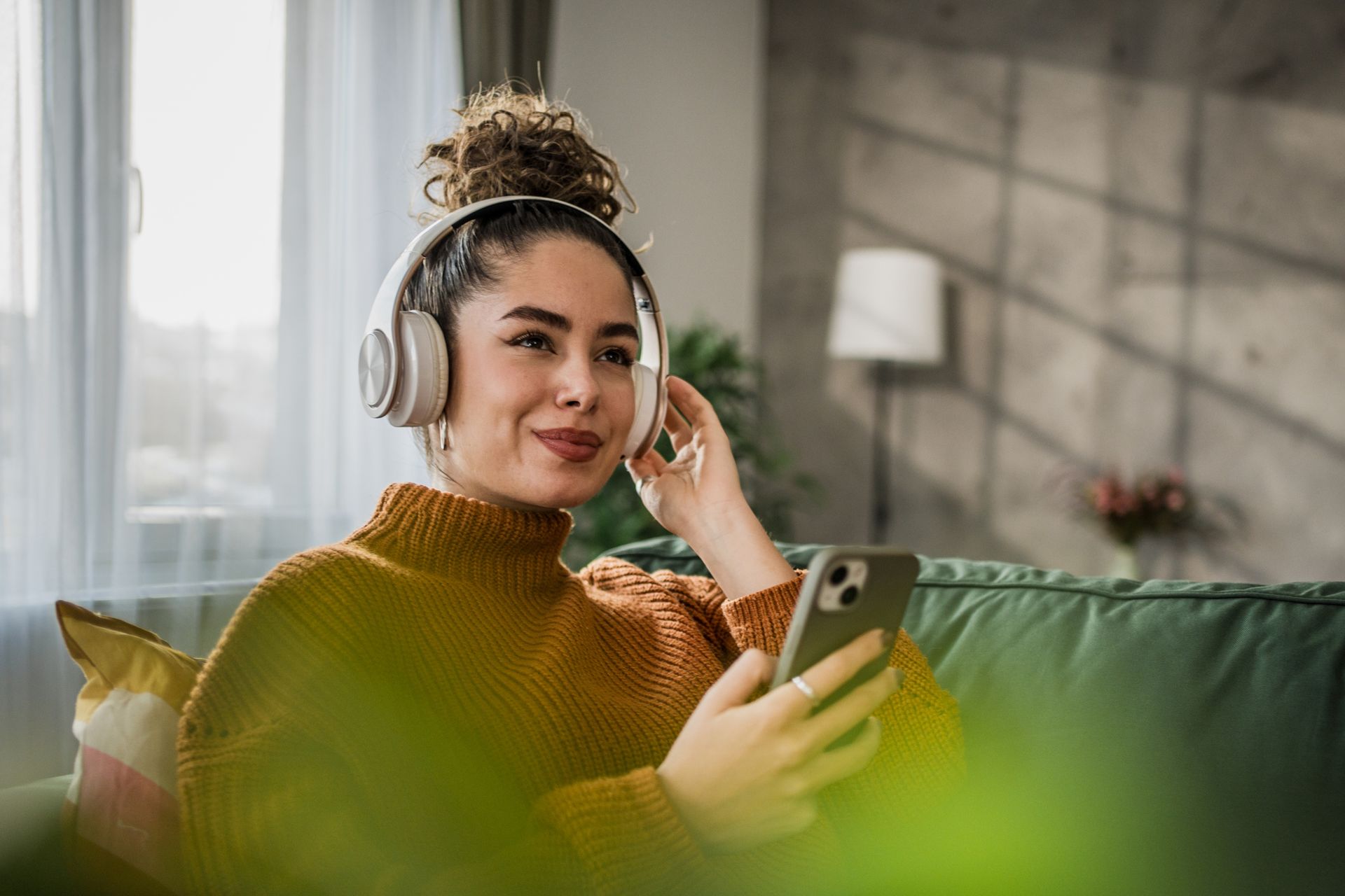 A woman is sitting on a couch wearing headphones and using a cell phone.