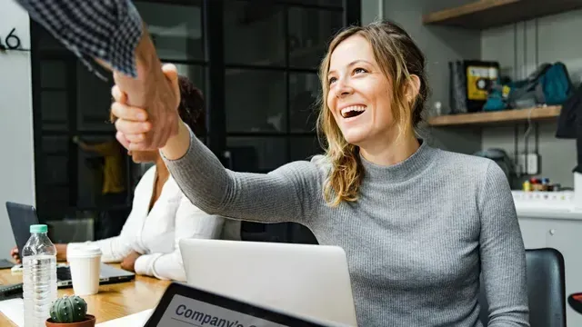 A woman is shaking hands with a man while sitting at a table with a laptop.