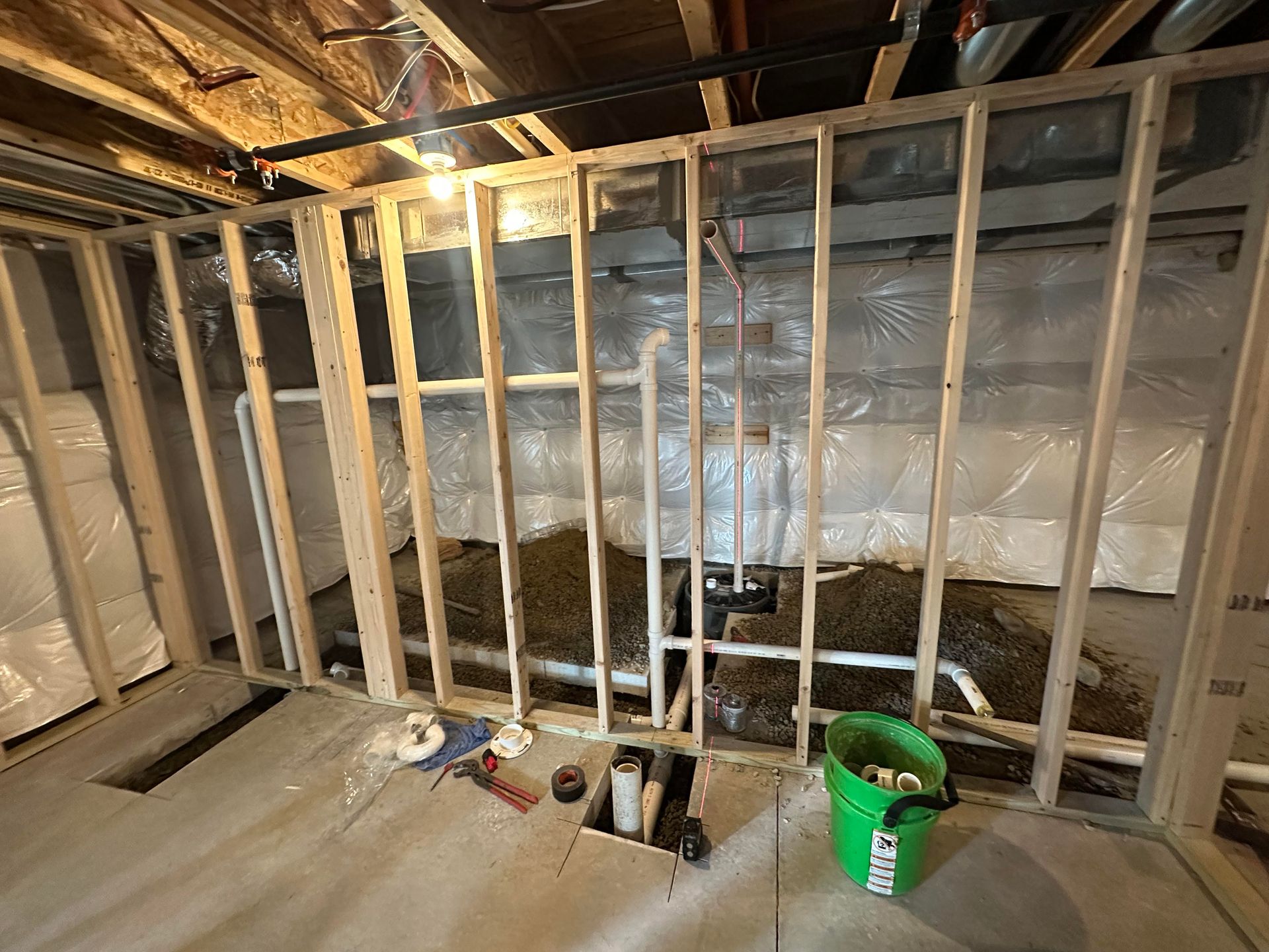 Interior view of a basement framing project. Wooden studs surround pipes and a green bucket sits on the concrete floor.