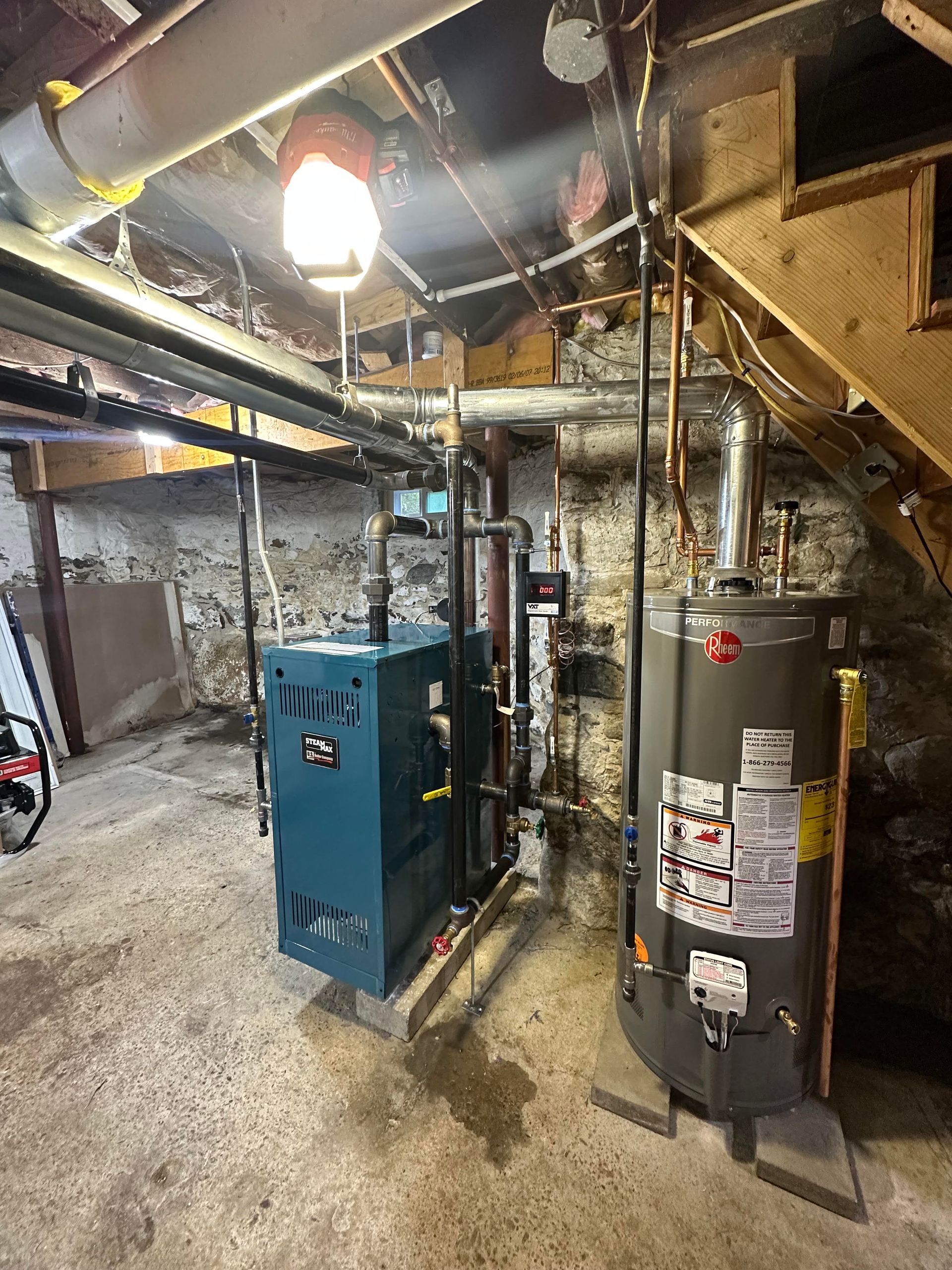 Basement with blue boiler, water heater, pipes, under stairs. Lighting is on.