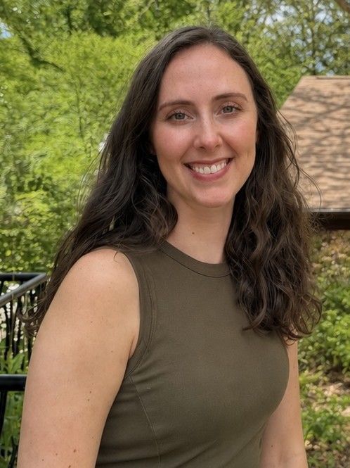 Smiling woman in an olive sleeveless top standing outdoors by trees and a railing
