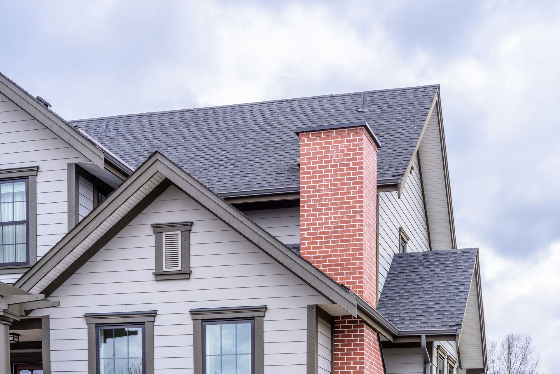 Suburban house roof and gables with gray siding and a red brick chimney under a cloudy sky