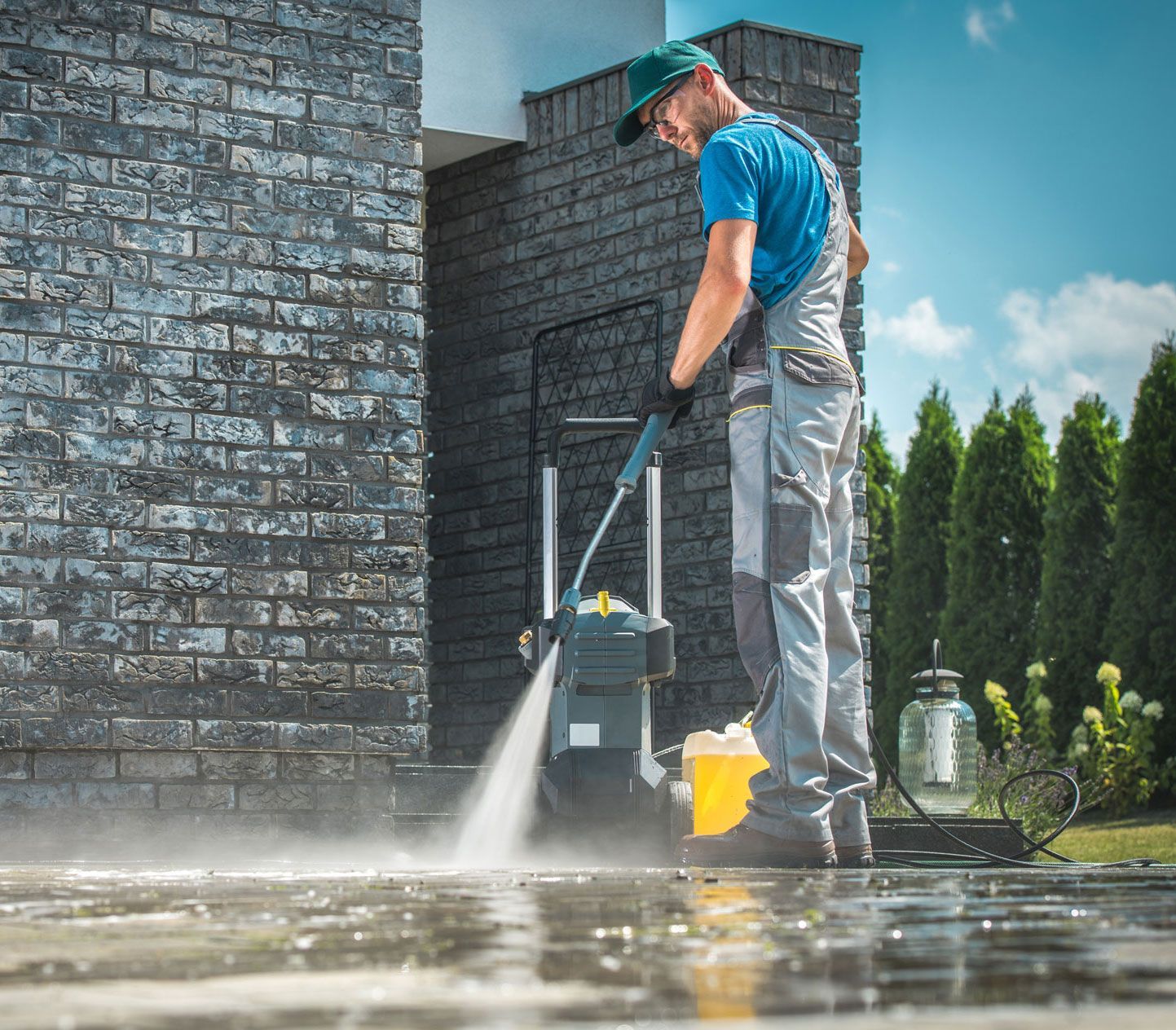 Worker pressure-washing a brick wall and wet patio beside a house, wearing boots and gloves.