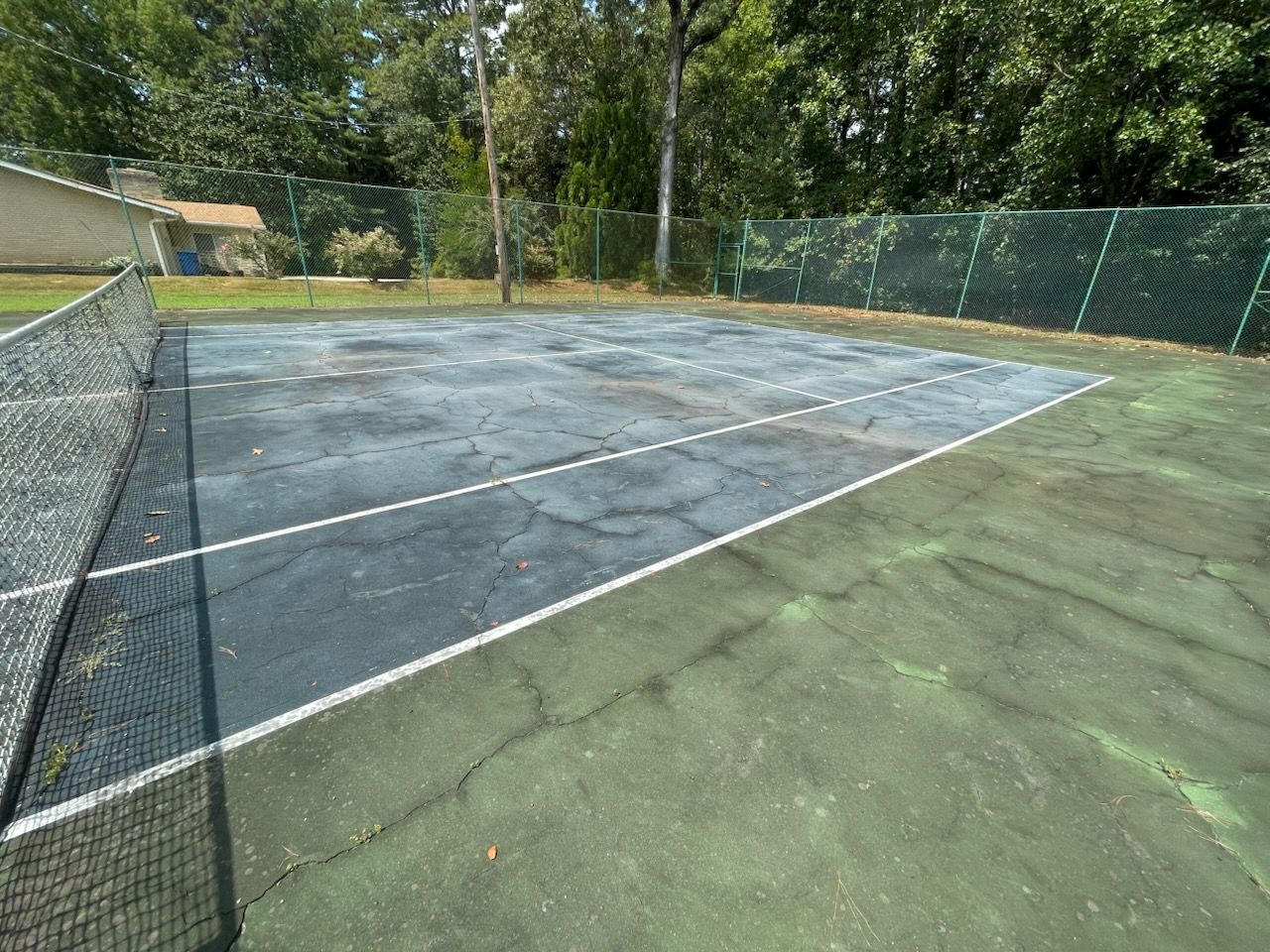 Empty outdoor tennis court with worn blue surface, green sideline area, and trees in the background
