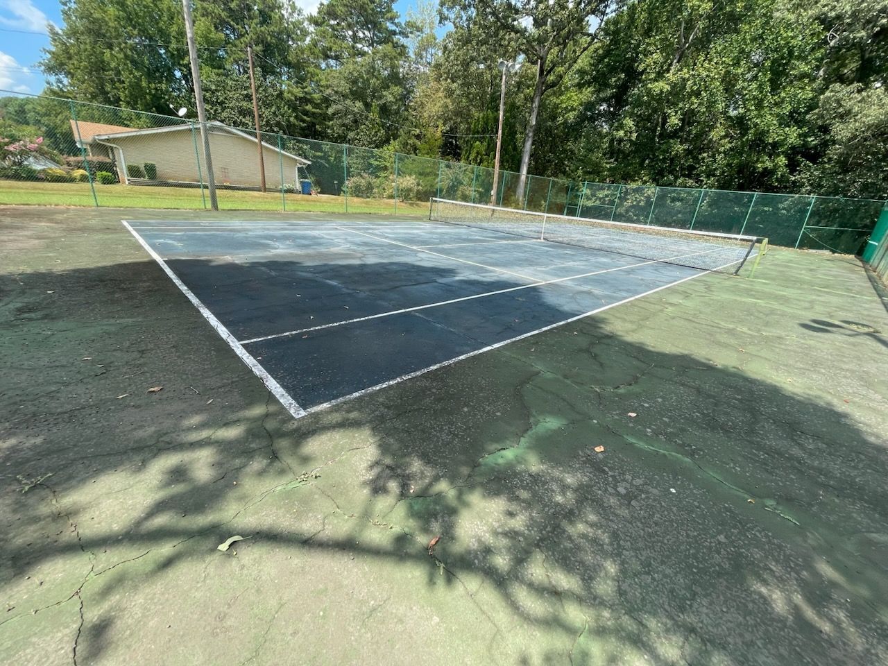Outdoor tennis court with dark surface and white lines, surrounded by grass and trees in sunlight
