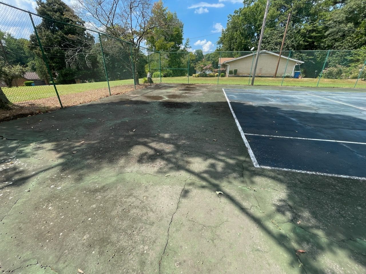 Outdoor tennis court with chain-link fence, trees, and a small building in the background.