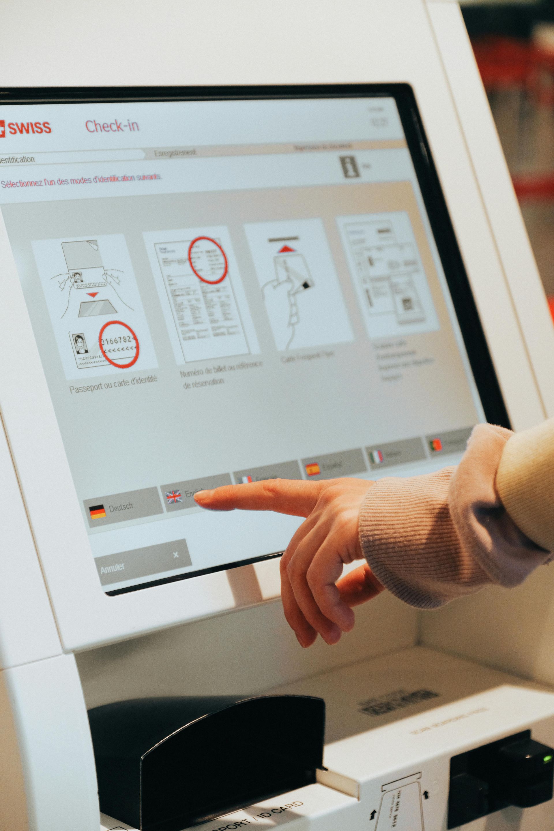 Person's finger touching a touchscreen at an airport kiosk, selecting language option.