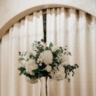 White floral arrangement on a stand before cream drapery and an arched backdrop