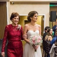 Bride in a white dress holds a pink bouquet beside an older woman in a red dress at an indoor event