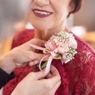 Hands pin a pink floral corsage onto a red lace dress while a smiling person looks on