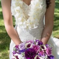 Bride holding purple and white bouquet in a garden, wearing a white floral dress