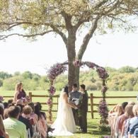 Outdoor wedding ceremony under a tree with floral arch, bride and groom exchanging vows