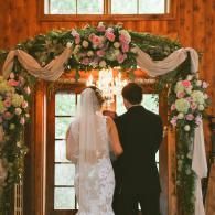 Bride and groom standing beneath a floral wedding arch in a rustic wooden venue