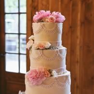 Three-tier white wedding cake with pink flowers and lace details by a window