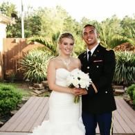 Bride and groom posing outdoors in wedding attire, with greenery and a wooden walkway behind them