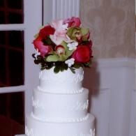 White tiered wedding cake topped with pink and red roses beside a window