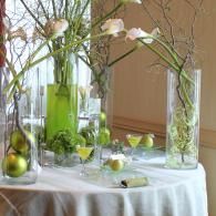 Glass vases with pale flowers and green fruit on a white tablecloth